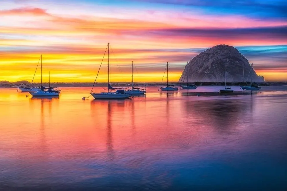Sail boats sitting in water during sunset with a single mountain in the background